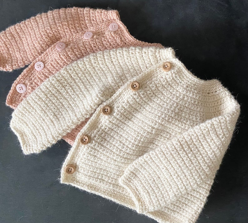 May include: Three handmade crochet baby cardigans in soft colors. The top cardigan is a light pink, the middle one is off-white, and the bottom one is a creamy white. Each cardigan has wooden buttons and a ribbed texture.