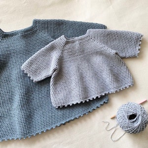 May include: Two crocheted sweaters in different shades of blue. The smaller sweater is light blue and has short sleeves. The larger sweater is a darker blue and has long sleeves. Both sweaters have a scalloped edge.