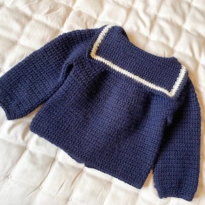May include: A navy blue crocheted sweater with a white trim around the neckline. The sweater has a sailor-style collar.