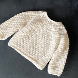 May include: Cream-colored, hand-crocheted sweater with long sleeves. The sweater has a round neckline and a textured, ribbed pattern throughout. The yarn appears to be a soft, natural fiber. A cozy, handmade garment.