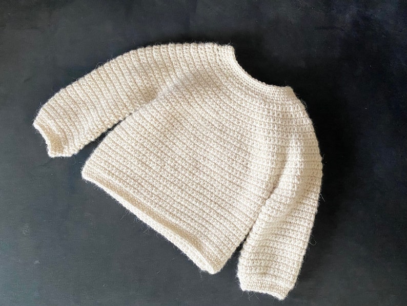 May include: Cream-colored, hand-crocheted sweater with long sleeves. The sweater has a round neckline and a textured, ribbed pattern throughout. The yarn appears to be a soft, natural fiber. A cozy, handmade garment.