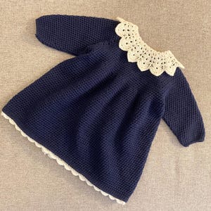 Puede incluir: Un vestido de punto azul marino con cuello y ribete de ganchillo blanco. El vestido tiene mangas largas y un corte holgado.