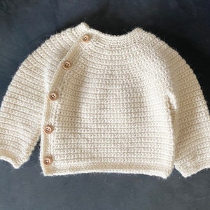 May include: Cream-colored, hand-crocheted baby cardigan with long sleeves and wooden buttons. The sweater features a round neckline and a button closure on the left side. The yarn has a textured appearance, and the buttons are light brown.