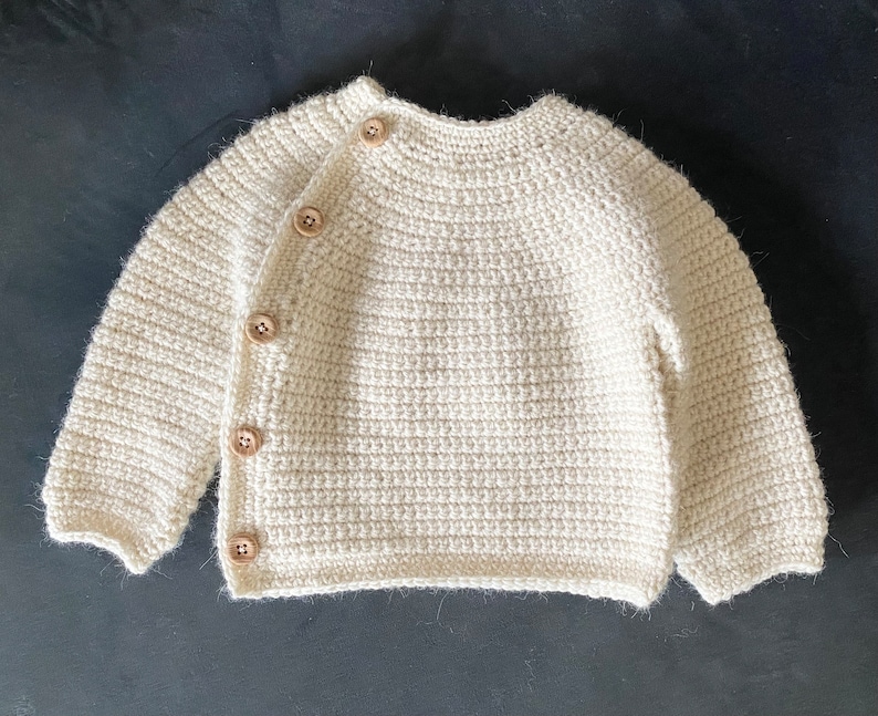 May include: Cream-colored, hand-crocheted baby cardigan with long sleeves and wooden buttons. The sweater features a round neckline and a button closure on the left side. The yarn has a textured appearance, and the buttons are light brown.
