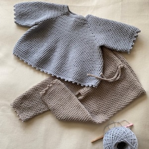 May include: A set of crocheted baby clothes in light blue and brown. The top has a round neckline and 3/4 sleeves. The pants have a straight leg and a cuffed hem. The yarn is in a skein next to the clothes.