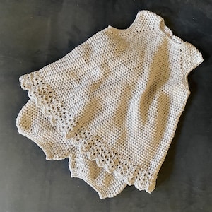 以下が含まれることがあります： 明るいベージュのかぎ針編みのトップスとショーツのセット。トップスはハイネックでキャップスリーブです。ショーツはスカラップエッジです。セットは、質感のあるオープンウィーブ生地で作られています。