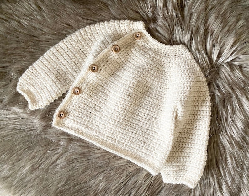 May include: Cream-colored, crocheted baby cardigan with four wooden buttons. The long-sleeved sweater is handmade with a textured stitch and is displayed on a fluffy, gray surface. A cozy and stylish garment for a baby.
