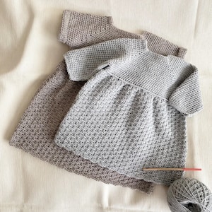 Puede incluir: Tres vestidos de ganchillo hechos a mano en tonos grises y taupe se muestran sobre una superficie color crema. Los vestidos tienen un patrón de puntada texturizado y un diseño sencillo. También se ven un ovillo de lana gris y un ganchillo de madera.