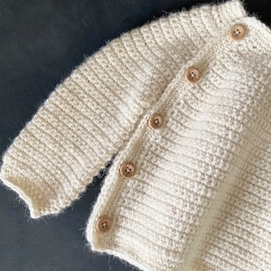 May include: Cream-colored, hand-knitted cardigan with wooden buttons. The textured knitwear features a ribbed collar and cuffs, and the buttons are evenly spaced down the front. The soft, neutral color makes it a versatile piece for layering.