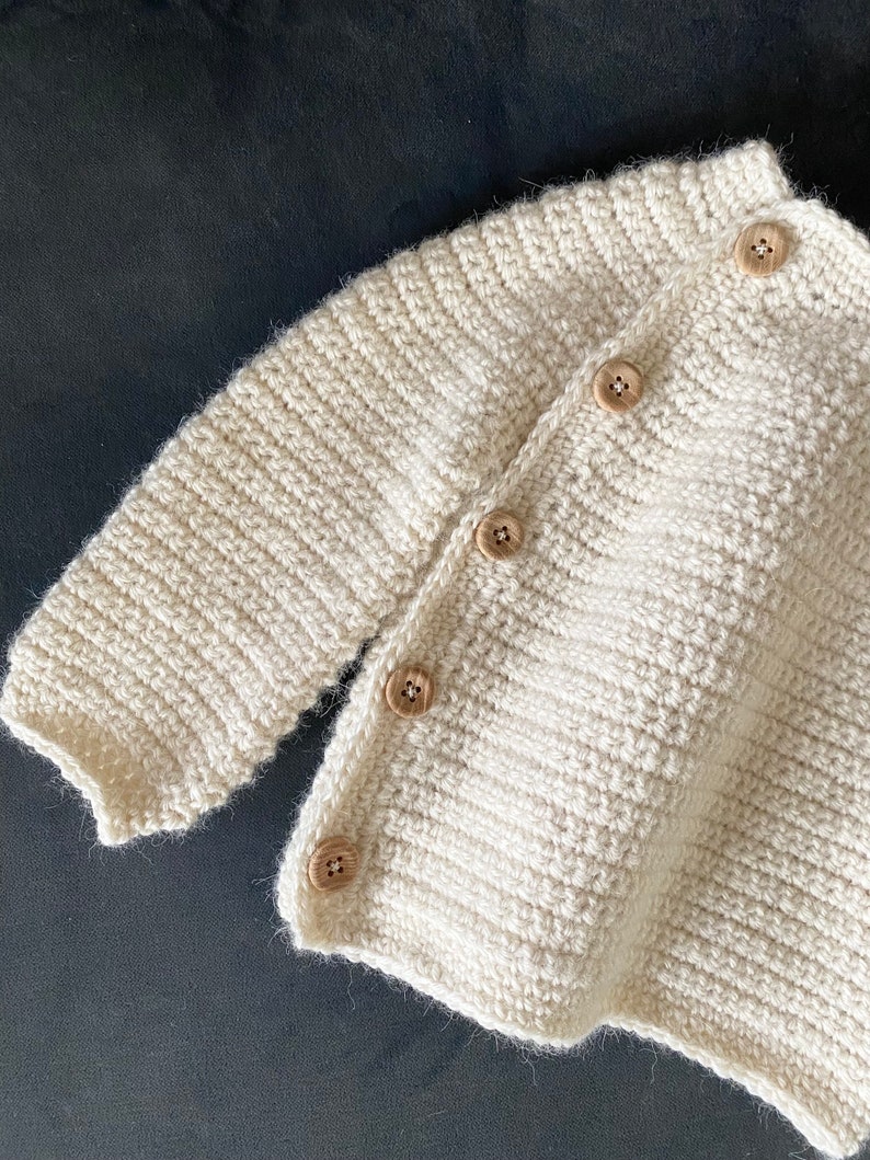 May include: Cream-colored, hand-knitted cardigan with wooden buttons. The textured knitwear features a ribbed collar and cuffs, and the buttons are evenly spaced down the front. The soft, neutral color makes it a versatile piece for layering.