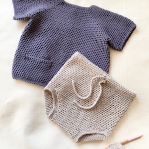 Puede incluir: Un conjunto de ropa de bebé tejido a crochet con una camiseta de manga corta de color morado oscuro y pantalones cortos beige. La camiseta tiene un bolsillo en la parte delantera. También se ve una bola de hilo beige y un gancho de crochet en la imagen.