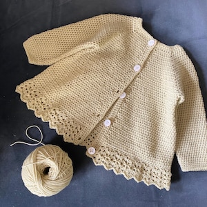 以下が含まれることがあります： 長袖と白いボタンが付いた、薄いベージュのかぎ針編みのカーディガン。下端には装飾的なスカラップトリムが付いています。お揃いの糸玉がセーターの隣にあり、手作りのアイテムであることを示唆しています。カーディガンはおそらく赤ちゃんまたは幼児用です。