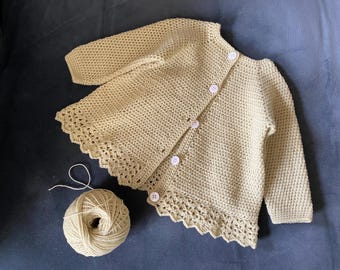 Peut inclure: Un cardigan au crochet beige clair, fait à la main, avec des manches longues et des boutons blancs. Le cardigan a un bord festonné et est présenté avec une pelote de fil assortie. Le vêtement est probablement pour un bébé ou un tout-petit.