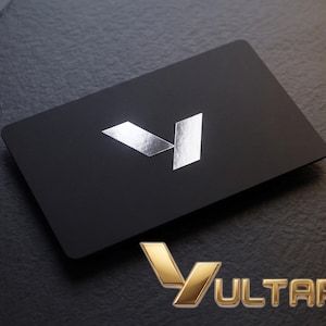May include: A rectangular black card with rounded corners featuring a centered silver metallic V-shaped logo. Below the card, the word VULTAP is displayed in a three-dimensional gold metallic font with a matching V-shaped icon to the left.