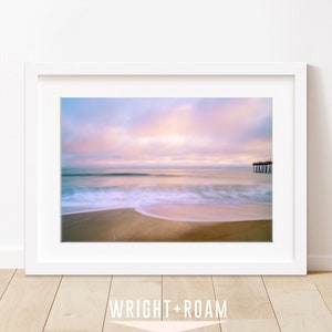 May include: A framed photograph of a beach scene with a pier in the distance. The sky is a soft pink and purple, and the waves are crashing on the shore. The sand is a light brown color.