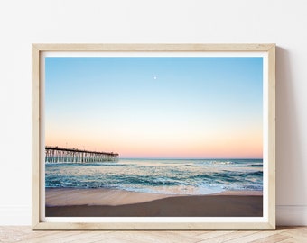 May include: A framed photograph of a wooden pier extending into the ocean at sunset. The sky is a gradient of blue, pink, and orange. The waves are crashing on the sandy beach.