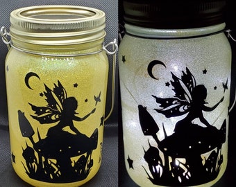 May include: A glass jar with a gold lid and a fairy silhouette design. The jar is filled with glitter and has a wire handle. The fairy is sitting on a mushroom with stars and a crescent moon in the background.