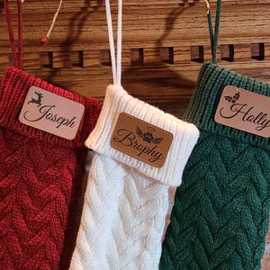 May include: Three cable-knit Christmas stockings in red, white, and green hang from a wooden mantel. Each stocking has a leather patch with a name: "Joseph", "Brophy", and "Holly". The stockings are decorated with a deer, angel wings, and holly.