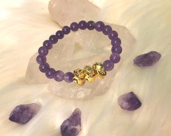 May include: A purple amethyst bead bracelet with three gold-colored accent beads. The bracelet is surrounded by raw amethyst crystals and rests on a white, fluffy surface. The beads are a deep, rich purple color.