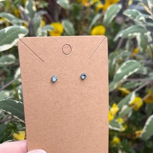 May include: A pair of small, round, light blue stud earrings mounted on a rectangular brown cardboard display card. The earrings feature a single faceted stone set in a simple metal prong setting, centered on the card.