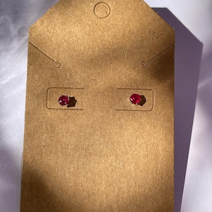 May include: A pair of round, faceted, deep red stud earrings secured to a rectangular brown cardboard display card. The earrings feature a brilliant cut and are mounted in a simple metal setting, presenting a classic and symmetrical appearance.