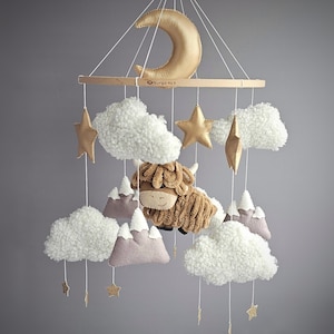 May include: A baby mobile featuring a gold crescent moon, stars, fluffy white clouds, and a brown Highland cow. The mobile includes light brown mountains and is suspended from a wooden ring.