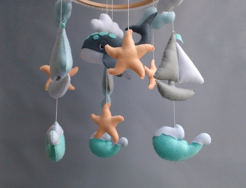 Puede incluir: Un m&oacute;vil para beb&eacute; con tem&aacute;tica n&aacute;utica. Presenta una ballena, un velero, una estrella de mar y nubes, todo hecho de fieltro en tonos azules, verdes y blancos.