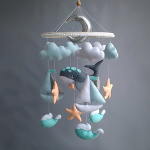 Puede incluir: Un m&oacute;vil para beb&eacute; con una ballena, nubes, veleros y estrellas de mar. El m&oacute;vil est&aacute; hecho de fieltro y tiene una luna creciente plateada en la parte superior.