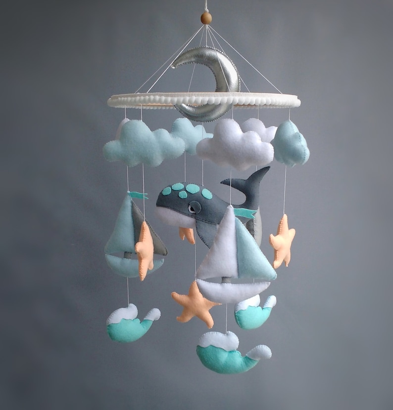Puede incluir: Un m&oacute;vil para beb&eacute; con una ballena, nubes, veleros y estrellas de mar. El m&oacute;vil est&aacute; hecho de fieltro y tiene una luna creciente plateada en la parte superior.