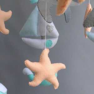 Puede incluir: Un m&oacute;vil para beb&eacute; con un velero, estrellas de mar y gaviotas hechas de fieltro. El velero es azul claro y blanco con un acento verde. Las estrellas de mar son melocot&oacute;n y las gaviotas son grises.