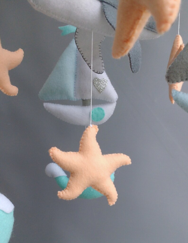 Puede incluir: Un m&oacute;vil para beb&eacute; con un velero, estrellas de mar y gaviotas hechas de fieltro. El velero es azul claro y blanco con un acento verde. Las estrellas de mar son melocot&oacute;n y las gaviotas son grises.