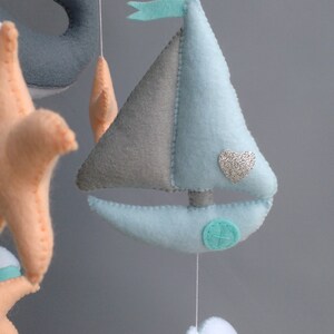 Puede incluir: Un m&oacute;vil para beb&eacute; con un velero, una ballena, una estrella de mar y nubes. El velero es azul claro y gris con un coraz&oacute;n de purpurina plateada. La ballena es gris, la estrella de mar es naranja y las nubes son azul claro.