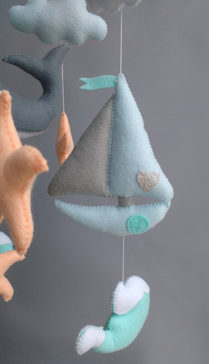 Puede incluir: Un m&oacute;vil para beb&eacute; con un velero, una ballena, una estrella de mar y nubes. El velero es azul claro y gris con un coraz&oacute;n de purpurina plateada. La ballena es gris, la estrella de mar es naranja y las nubes son azul claro.