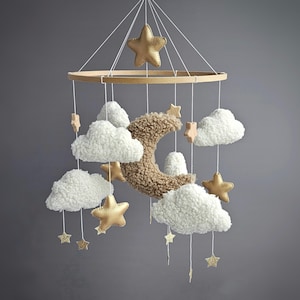 Może przedstawiać: Karuzela dla dziecka z drewnianym pierścieniem, białymi pluszowymi chmurkami, złotymi gwiazdami i brązowym księżycem w kształcie półksiężyca. Gwiazdy są złote, a karuzela zawieszona jest na białych sznurkach. Chmury są miękkie.