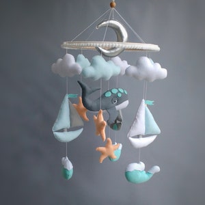 Puede incluir: Un m&oacute;vil para beb&eacute; con una ballena gris, dos veleros blancos, dos nubes, una luna creciente y cuatro estrellas de mar naranjas. El m&oacute;vil est&aacute; hecho de fieltro y est&aacute; colgado de un anillo de madera.