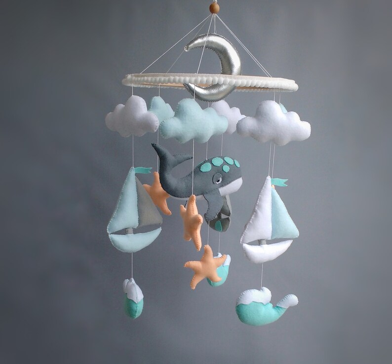 Puede incluir: Un m&oacute;vil para beb&eacute; con una ballena gris, dos veleros blancos, dos nubes, una luna creciente y cuatro estrellas de mar naranjas. El m&oacute;vil est&aacute; hecho de fieltro y est&aacute; colgado de un anillo de madera.