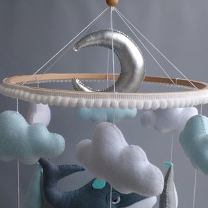 Puede incluir: Un m&oacute;vil para beb&eacute; con un aro de madera, ribete de pompones blancos y nubes de fieltro colgantes, una ballena y una luna creciente plateada.