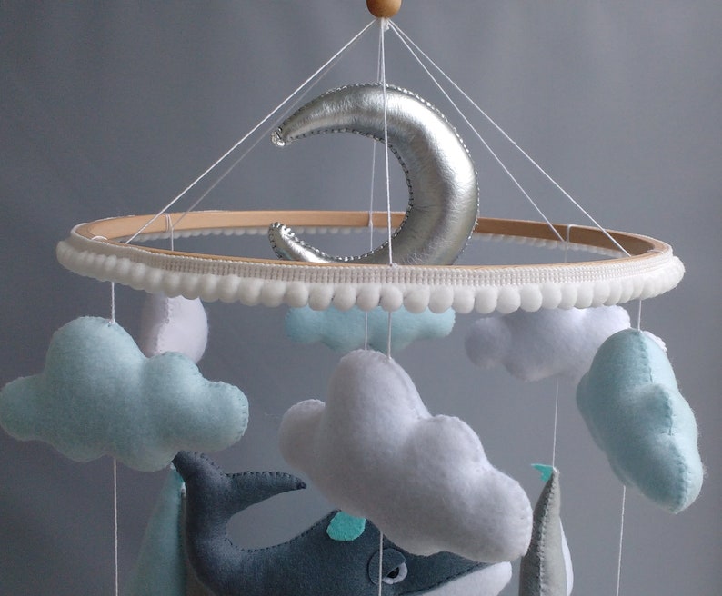 Puede incluir: Un m&oacute;vil para beb&eacute; con un aro de madera, ribete de pompones blancos y nubes de fieltro colgantes, una ballena y una luna creciente plateada.