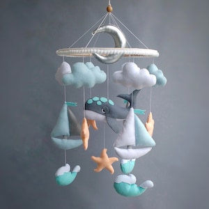 Puede incluir: Un m&oacute;vil para beb&eacute; con una ballena gris, dos veleros, dos nubes, una estrella de mar y una luna creciente. El m&oacute;vil est&aacute; hecho de fieltro y tiene un esquema de color azul claro, blanco y gris.