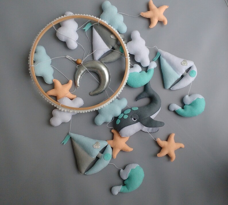 Puede incluir: Un m&oacute;vil para beb&eacute; con tem&aacute;tica n&aacute;utica. Presenta una ballena, un velero, nubes, estrellas y una luna creciente. El m&oacute;vil est&aacute; hecho de fieltro y est&aacute; colgado de un aro de madera.