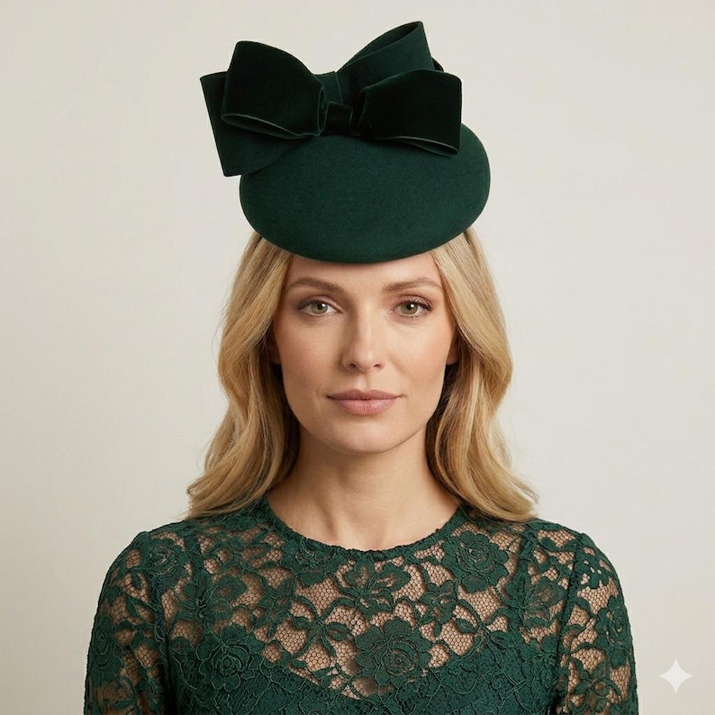K&ouml;nnte beinhalten: Ein dunkelgr&uuml;ner Hut mit einer gro&szlig;en Samtschleife. Der Hut ist im Pillbox-Stil gehalten, mit einer abgerundeten Krone und einer breiten, dekorativen Schleife. Die Schleife ist aus dunklerem gr&uuml;nen Samt gefertigt. Der Hut wird von einer Person mit blonden Haaren getragen.