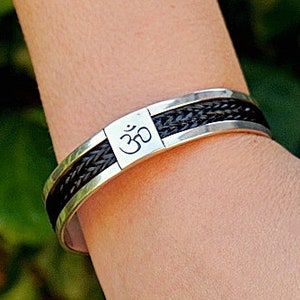 May include: A silver bracelet with a black braided leather band and an Om symbol engraved on the front.