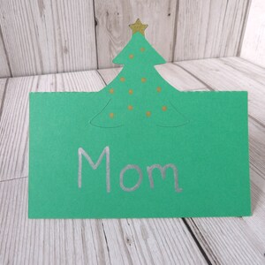 A green place card with a Christmas tree cut out of the top. The tree has a gold star and old dots for baubles on it. Mom is written on the card as an example.