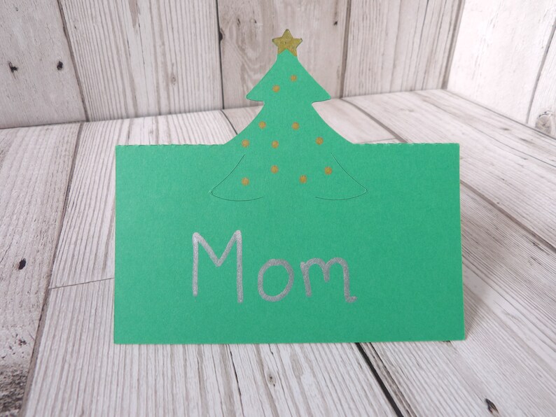 A green place card with a Christmas tree cut out of the top. The tree has a gold star and old dots for baubles on it. Mom is written on the card as an example.