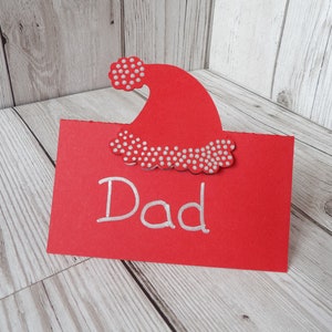 A red place card with a Santa hat cut out of the top. The hat has silver dots on the bobble and trim. Dad is written on the card as an example of it in use.