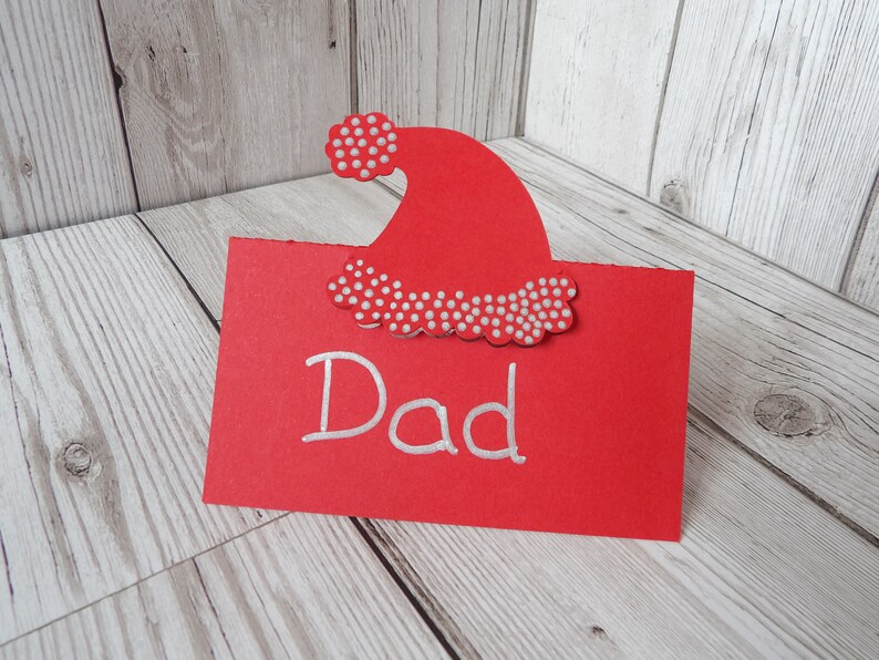 A red place card with a Santa hat cut out of the top. The hat has silver dots on the bobble and trim. Dad is written on the card as an example of it in use.