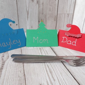 A blue place card with an elf hat cut out of the top, a green place card with a Christmas tree cut out of the top and a red place card with a Santa hat cut out of the top. In front are a knife and fork for comparison of size.