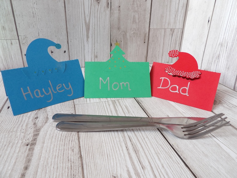 A blue place card with an elf hat cut out of the top, a green place card with a Christmas tree cut out of the top and a red place card with a Santa hat cut out of the top. In front are a knife and fork for comparison of size.