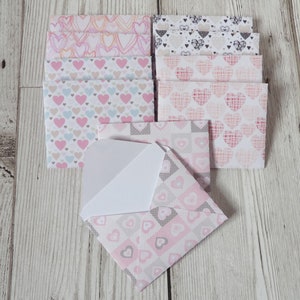 May include: A set of eight small envelopes with different pink and white heart patterns. The envelopes are folded and stacked on a light wood surface.