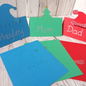 A blue place card with an elf hat cut out of the top, a green place card with a Christmas tree cut out of the top and a red place card with a Santa hat cut out of the top. The 3 place cards are also shown flat in front.